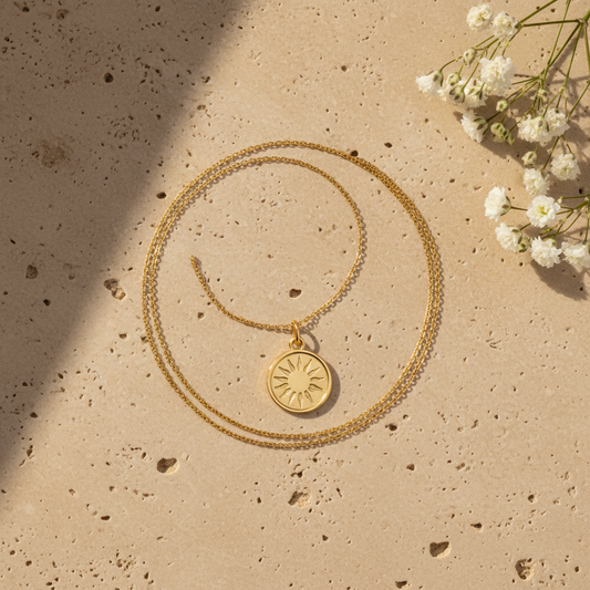 Gold coin pendant necklace on warm stone surface with flowers